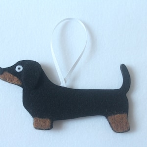 May include: A felt dachshund ornament. The dog is black with brown paws and snout. It has a white ribbon for hanging. The dog's eye is white with a black center. The ornament is on a white background.