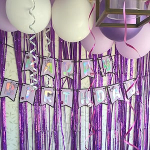 May include: A Happy Birthday banner with light purple pennants and iridescent letters, featuring small character icons and bows. The banner is displayed against a backdrop of metallic purple fringe curtains, accented by white and purple balloons.