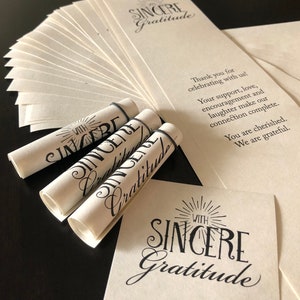 May include: Three rolled up cards with the text "With Sincere Gratitude" printed on them. The cards are on a table with a larger card with the same text in the background.