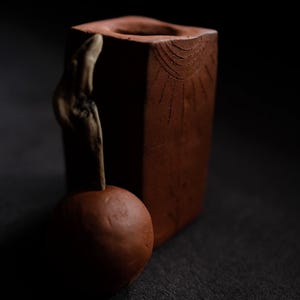 May include: A brown, square wooden vase with a natural wood accent and a round, brown object in front. The vase has a carved sunburst design. The objects are set against a dark background.