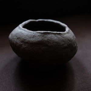 Puede incluir: Un cuenco de cerámica gris oscuro hecho a mano, con una superficie texturizada y una forma ligeramente irregular. El cuenco tiene una abertura ancha y una serie de líneas grabadas alrededor del exterior. El cuenco se muestra sobre una superficie oscura.