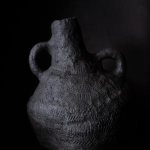 Puede incluir: Un jarrón de cerámica gris oscuro y texturizado con dos asas de bucle y un cuello estrecho. El jarrón tiene un cuerpo bulboso y una apariencia áspera, hecha a mano, sobre un fondo negro.