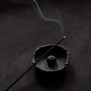 May include: A black incense stick with a glowing orange tip rests in a small, dark ceramic bowl. Wisps of white smoke curl upwards against a dark background, creating a calming and aromatic scene.