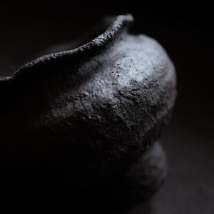 Puede incluir: Primer plano de un cuenco de cerámica gris oscuro y texturizado con un borde rugoso e irregular. El cuenco tiene un cuerpo redondeado y se asienta sobre una pequeña base circular. La iluminación crea sombras que enfatizan la textura.