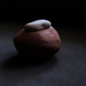 Puede incluir: Una pequeña escultura de arcilla redondeada, de color marrón rojizo, con una piedra gris oblonga más pequeña encima. Los objetos se colocan sobre un fondo oscuro, creando un contraste de texturas y colores. La escultura parece hecha a mano.