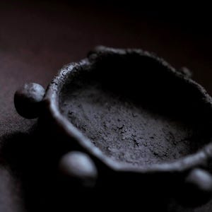 Puede incluir: Un objeto oscuro, texturizado, en forma de cuenco con tres acentos esféricos. El cuenco tiene una superficie rugosa y desigual y es de un color carbón profundo. El diseño general es minimalista y orgánico, lo que sugiere un artículo hecho a mano.