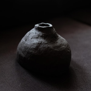 Puede incluir: Un jarrón de cerámica gris oscuro hecho a mano con una superficie texturizada. El jarrón tiene un cuerpo redondeado y una abertura estrecha en la parte superior. La forma general es orgánica y ligeramente irregular, lo que sugiere un origen artesanal.