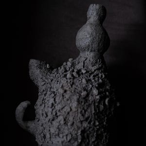 Puede incluir: Una escultura texturizada de color gris oscuro con una forma abstracta. La escultura tiene una superficie rugosa e irregular y una parte superior bulbosa. La forma general es orgánica e irregular, con un elemento curvo que se extiende desde el lateral. El fondo es negro sólido.