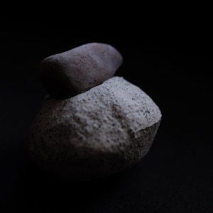 May include: Two stones stacked against a black backdrop. The lower stone is light grey and textured, while the upper stone is smaller and a darker grey. The lighting creates shadows, highlighting the shapes and textures of the stones.