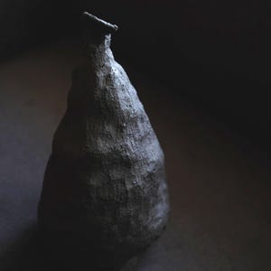 Puede incluir: Una escultura de forma cónica de color gris oscuro con una superficie rugosa y texturizada. La obra de arte tiene una parte superior pequeña y rectangular y una base redondeada. La escultura se encuentra sobre un fondo oscuro, creando un contraste dramático.