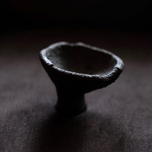 Puede incluir: Un cuenco de cerámica oscuro, texturizado y hecho a mano con una base de pedestal. El cuenco tiene una parte superior ancha y abierta y un borde áspero e irregular. La pieza es de color oscuro y mate, y se asienta sobre una superficie oscura.