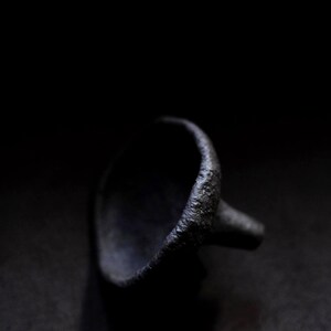 Puede incluir: Un anillo negro oscuro y texturizado con una parte superior redondeada y una pequeña proyección cilíndrica. La superficie del anillo parece rugosa y desigual, con un acabado mate. El fondo es negro sólido.