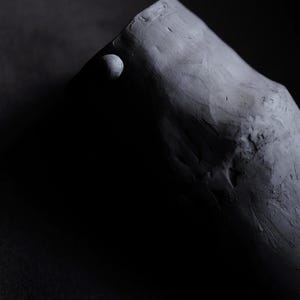 Puede incluir: Fotografía oscura de una escultura gris y texturizada. La escultura tiene una forma redondeada, similar a una montaña, con un pequeño detalle esférico en un lado. La iluminación crea sombras profundas.