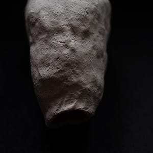 Puede incluir: Una escultura abstracta de color gris claro, de textura rugosa, con una parte superior redondeada y una base ensanchada. El objeto se encuentra sobre un fondo negro, resaltando su forma y los detalles de la superficie. La escultura recuerda a un recipiente o forma orgánica.