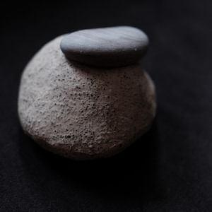 Op de afbeelding: Een getextureerde, lichtgrijze steen met een kleinere, gladde, donkergrijze steen die erop rust. De stenen staan ​​tegen een donkere achtergrond, wat een minimalistische en natuurlijke esthetiek creëert. De afbeelding benadrukt het contrast in texturen en tinten.