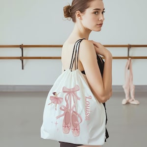May include: White drawstring bag featuring a pink ballerina, ballet shoes, and the name "Louisa" in pink script. The bag has black drawstrings and a pink bow design. The bag is designed for ballet.
