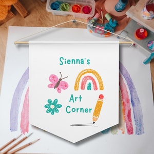 May include: A white banner with the text "Sienna's Art Corner" and illustrations of a butterfly, rainbow, flower, and pencil. The banner is suspended from a wooden rod.