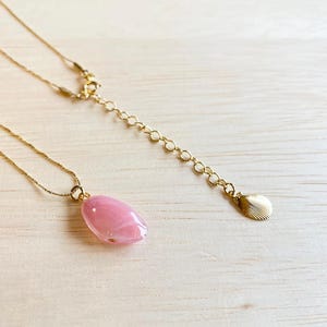 以下が含まれることがあります： ピンク色の楕円形ペンダントが付いた繊細なゴールドネックレス。ネックレスは細いチェーンと、留め金に小さな貝殻型のチャームが付いています。ペンダントはソフトピンク色です。