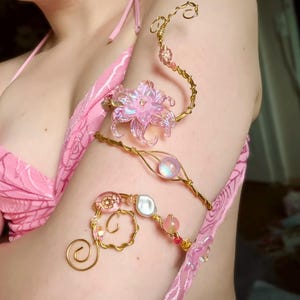 Könnte beinhalten: Ein Oberarmreif aus goldfarbenem Draht mit einer großen, irisierenden rosa Blume, einer zentralen kugelförmigen Perle sowie kleineren rosa und weißen Akzentperlen. Der Draht ist an beiden Enden spiralförmig gedreht.