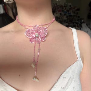 May include: A pink beaded necklace with a large flower pendant, a pearl centre, and dangling accents. The necklace is worn by a person wearing a white top. The flower pendant is the focal point of the necklace.