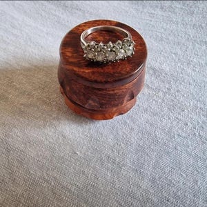 May include: A silver ring with multiple clear stones rests on a small, round, wooden box. The box is a rich, dark brown color with a carved design around the base. The ring is the focal point, showcasing its sparkling stones.