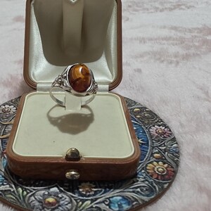 K&ouml;nnte beinhalten: Ein silberner Ring mit einem ovalen, bernsteinfarbenen Stein, pr&auml;sentiert in einer Vintage-Schmuckschatulle aus braunem Leder. Der Ring ist vor einem cremefarbenen Innenraum platziert. Die Schatulle steht auf einem dekorativen Untersetzer mit Blumenmuster.
