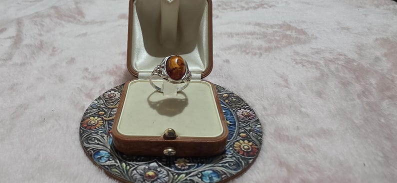 K&ouml;nnte beinhalten: Ein silberner Ring mit einem ovalen, bernsteinfarbenen Stein, pr&auml;sentiert in einer Vintage-Schmuckschatulle aus braunem Leder. Der Ring ist vor einem cremefarbenen Innenraum platziert. Die Schatulle steht auf einem dekorativen Untersetzer mit Blumenmuster.