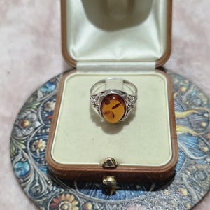 K&ouml;nnte beinhalten: Silberring mit einem ovalen Bernstein, pr&auml;sentiert in einer Vintage-Schmuckschatulle. Der Bernstein hat einen warmen, gold-orangenen Farbton. Der Ring ist in einem dekorativen Silberband gefasst.