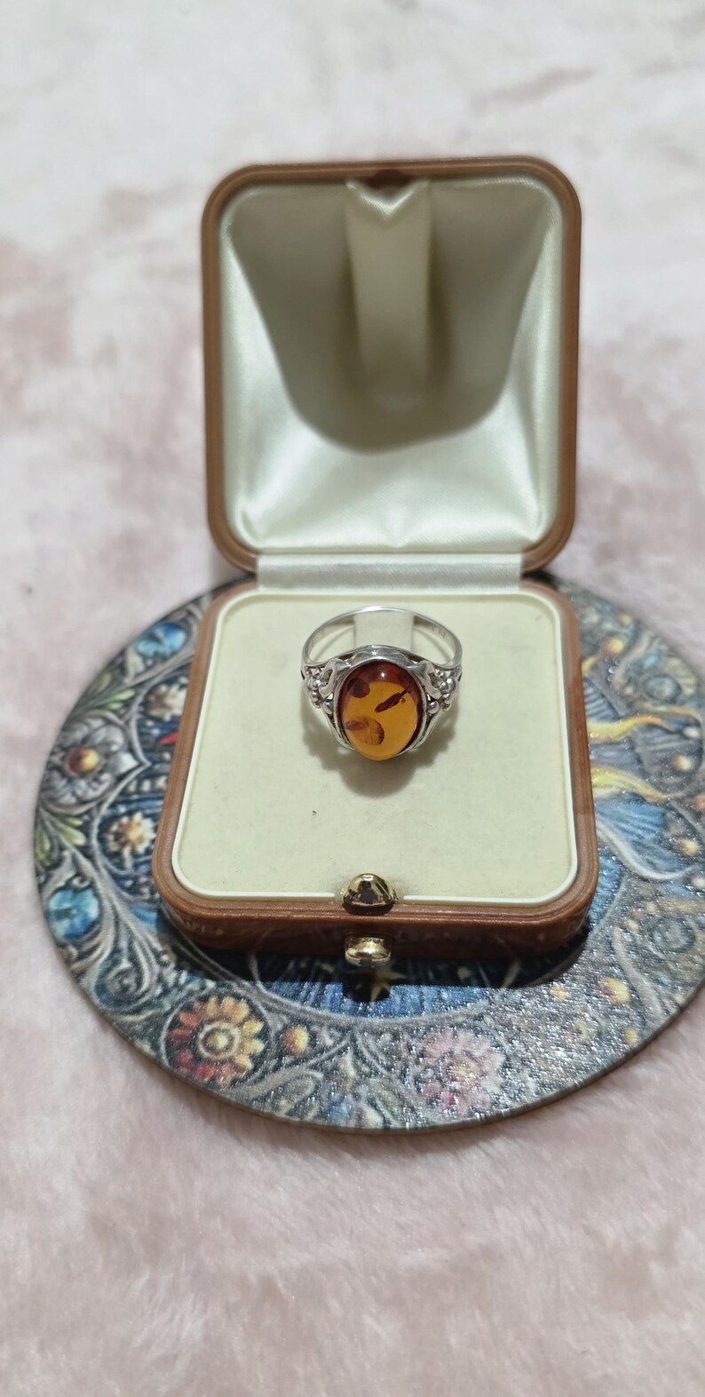 K&ouml;nnte beinhalten: Silberring mit einem ovalen Bernstein, pr&auml;sentiert in einer Vintage-Schmuckschatulle. Der Bernstein hat einen warmen, gold-orangenen Farbton. Der Ring ist in einem dekorativen Silberband gefasst.