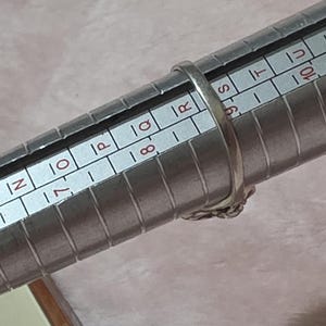 K&ouml;nnte beinhalten: Ein silberner Ring wird auf einem Ringma&szlig; gemessen. Der Ring befindet sich auf der Gr&ouml;&szlig;e 8. Der Ring hat ein dekoratives Design. Das Ringma&szlig; ist silberfarben und hat Messmarkierungen.
