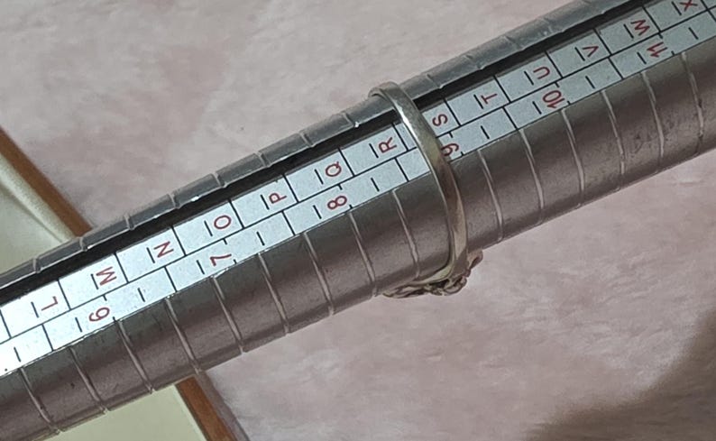 K&ouml;nnte beinhalten: Ein silberner Ring wird auf einem Ringma&szlig; gemessen. Der Ring befindet sich auf der Gr&ouml;&szlig;e 8. Der Ring hat ein dekoratives Design. Das Ringma&szlig; ist silberfarben und hat Messmarkierungen.