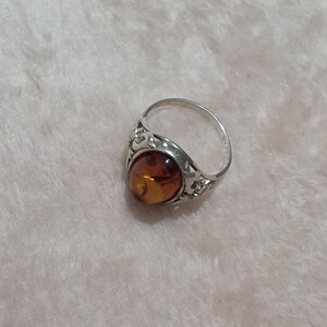 K&ouml;nnte beinhalten: Ein silberner Ring mit einem ovalen Bernstein. Der Ring hat ein verziertes, durchbrochenes Design um den Stein und am Band. Der Bernstein hat eine warme, durchscheinende orange Farbe. Der Ring ist vor einem weichen, rosa Hintergrund platziert.