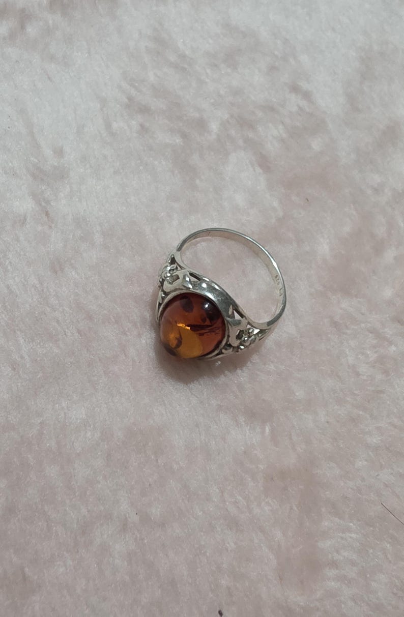 K&ouml;nnte beinhalten: Ein silberner Ring mit einem ovalen Bernstein. Der Ring hat ein verziertes, durchbrochenes Design um den Stein und am Band. Der Bernstein hat eine warme, durchscheinende orange Farbe. Der Ring ist vor einem weichen, rosa Hintergrund platziert.