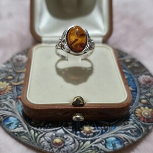 K&ouml;nnte beinhalten: Ein silberner Ring mit einem ovalen Bernstein, pr&auml;sentiert in einer Vintage-Schmuckschatulle aus braunem Leder. Der Ring hat verzierte Details. Die Schatulle liegt auf einer dekorativen, floral gemusterten Oberfl&auml;che.