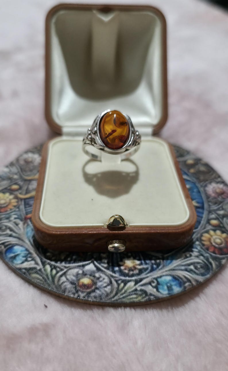 K&ouml;nnte beinhalten: Ein silberner Ring mit einem ovalen Bernstein, pr&auml;sentiert in einer Vintage-Schmuckschatulle aus braunem Leder. Der Ring hat verzierte Details. Die Schatulle liegt auf einer dekorativen, floral gemusterten Oberfl&auml;che.