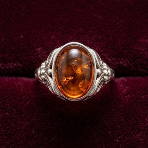 K&ouml;nnte beinhalten: Ein Silberring mit einem ovalen Bernstein-Edelstein. Der Bernstein ist ein warmes, durchscheinendes Orange mit sichtbaren Einschl&uuml;ssen. Der Ring hat ein Band mit dekorativen floralen und scrollartigen Details, was einen antiken oder Vintage-Look erzeugt.