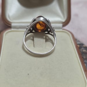 K&ouml;nnte beinhalten: Ein silberner Ring mit einem ovalen, bernsteinfarbenen Stein in einer dekorativen L&uuml;nette. Der Ring wird in einer Vintage-Schmuckschatulle mit cremefarbenem Innenraum und braunem &Auml;u&szlig;eren pr&auml;sentiert.