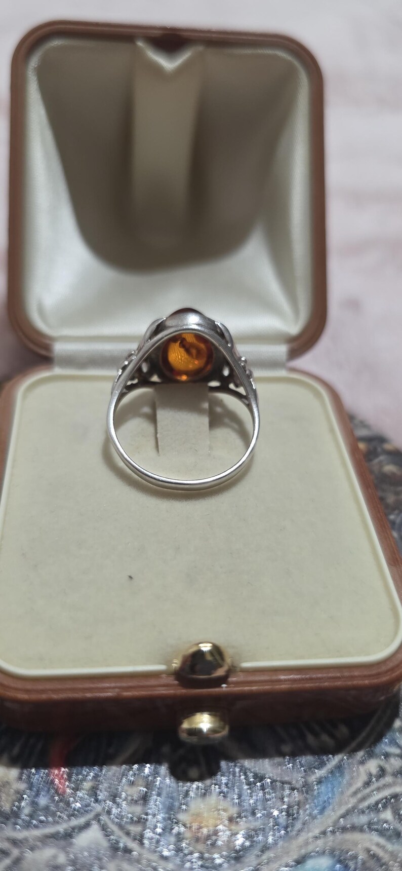 K&ouml;nnte beinhalten: Ein silberner Ring mit einem ovalen, bernsteinfarbenen Stein in einer dekorativen L&uuml;nette. Der Ring wird in einer Vintage-Schmuckschatulle mit cremefarbenem Innenraum und braunem &Auml;u&szlig;eren pr&auml;sentiert.