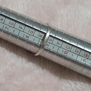 K&ouml;nnte beinhalten: Ein silberner Ring befindet sich auf einem Ringma&szlig;. Das Ringma&szlig; ist ein Metallzylinder mit aufgedruckten Ma&szlig;en und Buchstaben. Der Hintergrund ist ein weicher, hellrosa Stoff.