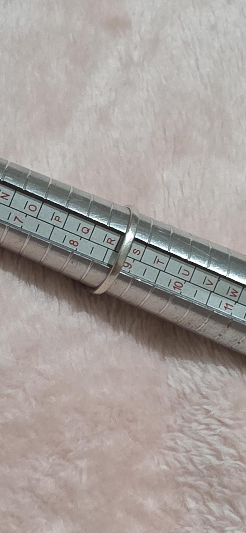 K&ouml;nnte beinhalten: Ein silberner Ring befindet sich auf einem Ringma&szlig;. Das Ringma&szlig; ist ein Metallzylinder mit aufgedruckten Ma&szlig;en und Buchstaben. Der Hintergrund ist ein weicher, hellrosa Stoff.