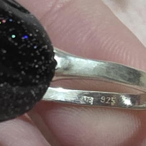 K&ouml;nnte beinhalten: Nahaufnahme eines Silberrings mit einem schwarzen, glitzernden Stein. Das Ringband ist mit "925" gestempelt. Der Stein hat blaue, gr&uuml;ne und rote Flecken. Der Ring wird in einer Hand gehalten.