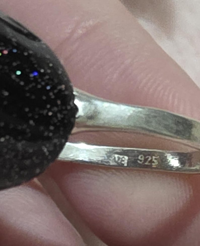 K&ouml;nnte beinhalten: Nahaufnahme eines Silberrings mit einem schwarzen, glitzernden Stein. Das Ringband ist mit "925" gestempelt. Der Stein hat blaue, gr&uuml;ne und rote Flecken. Der Ring wird in einer Hand gehalten.