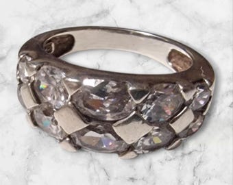Könnte beinhalten: Ein silberner Ring mit einem Band, das mit klaren, facettierten Steinen besetzt ist. Die Steine sind in einem sich wiederholenden Muster um den Umfang des Rings angeordnet. Der Ring ist vor einem Marmorhintergrund platziert.