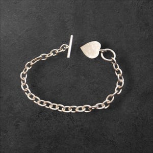 Puede incluir: Pulsera de cadena plateada con un dije en forma de corazón. La pulsera presenta un cierre de palanca y una serie de eslabones ovalados interconectados. El dije de corazón es plano y liso. La pulsera se muestra sobre una superficie oscura y texturizada.