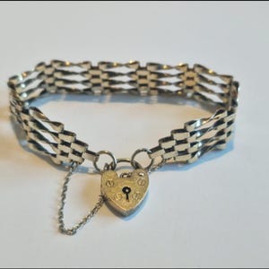 Puede incluir: Una pulsera plateada con un diseño tejido. La pulsera presenta un cierre de candado en forma de corazón con una pequeña cadena. La joya se exhibe sobre una superficie blanca.