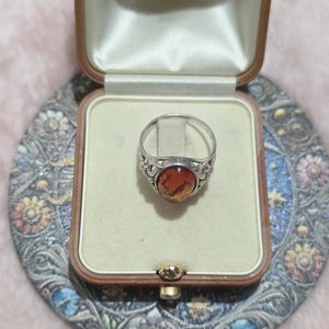 K&ouml;nnte beinhalten: Ein silberner Ring mit einem ovalen, bernsteinfarbenen Stein wird in einer Vintage-Schmuckschatulle pr&auml;sentiert. Der Ring hat ein detailliertes, verziertes Design um den Stein. Die Schatulle ist ge&ouml;ffnet und zeigt einen cremefarbenen Innenraum.