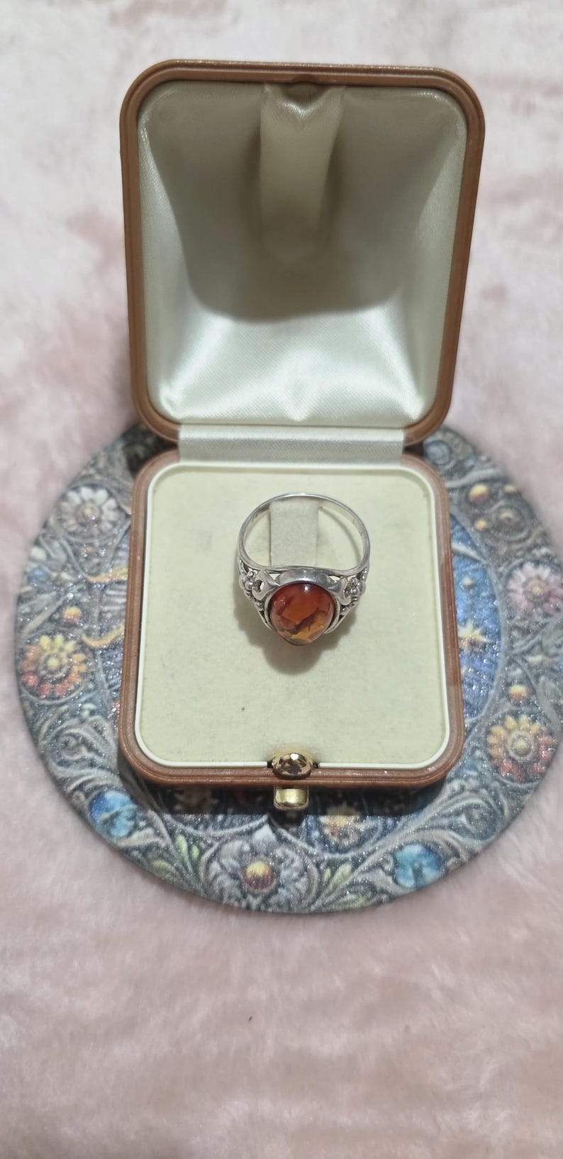 K&ouml;nnte beinhalten: Ein silberner Ring mit einem ovalen, bernsteinfarbenen Stein wird in einer Vintage-Schmuckschatulle pr&auml;sentiert. Der Ring hat ein detailliertes, verziertes Design um den Stein. Die Schatulle ist ge&ouml;ffnet und zeigt einen cremefarbenen Innenraum.