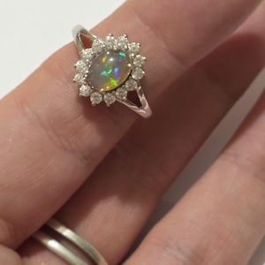 May include: A silver ring featuring an oval opal gemstone surrounded by a halo of small, round diamonds. The opal displays a play of color with iridescent hues. The ring is set on a split shank band.