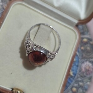 K&ouml;nnte beinhalten: Ein Silberring mit einem ovalen, bernsteinfarbenen Stein. Der Ring befindet sich in einer Vintage-Schmuckschatulle mit cremefarbenem Satinfutter. Der Ring hat ein verziertes Design um den Stein.