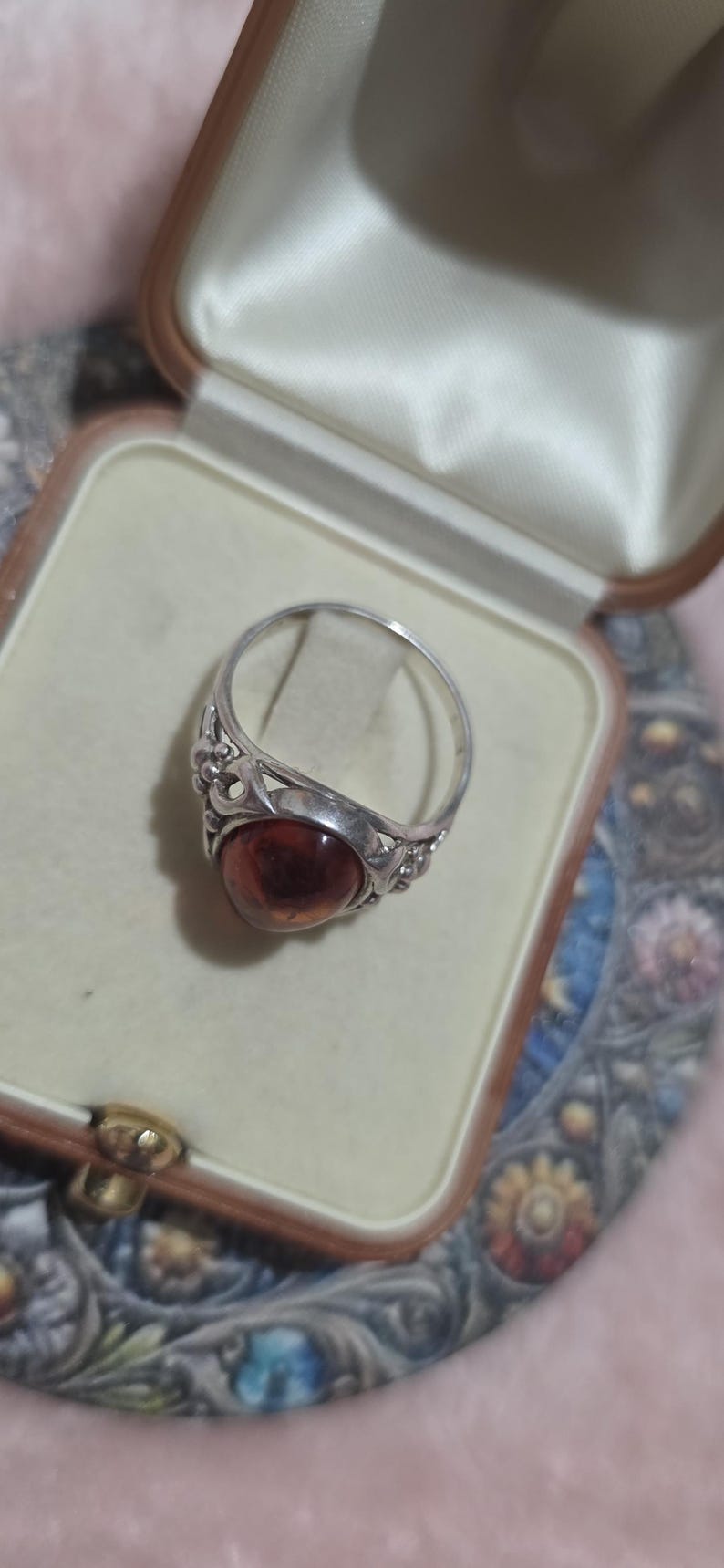 K&ouml;nnte beinhalten: Ein Silberring mit einem ovalen, bernsteinfarbenen Stein. Der Ring befindet sich in einer Vintage-Schmuckschatulle mit cremefarbenem Satinfutter. Der Ring hat ein verziertes Design um den Stein.